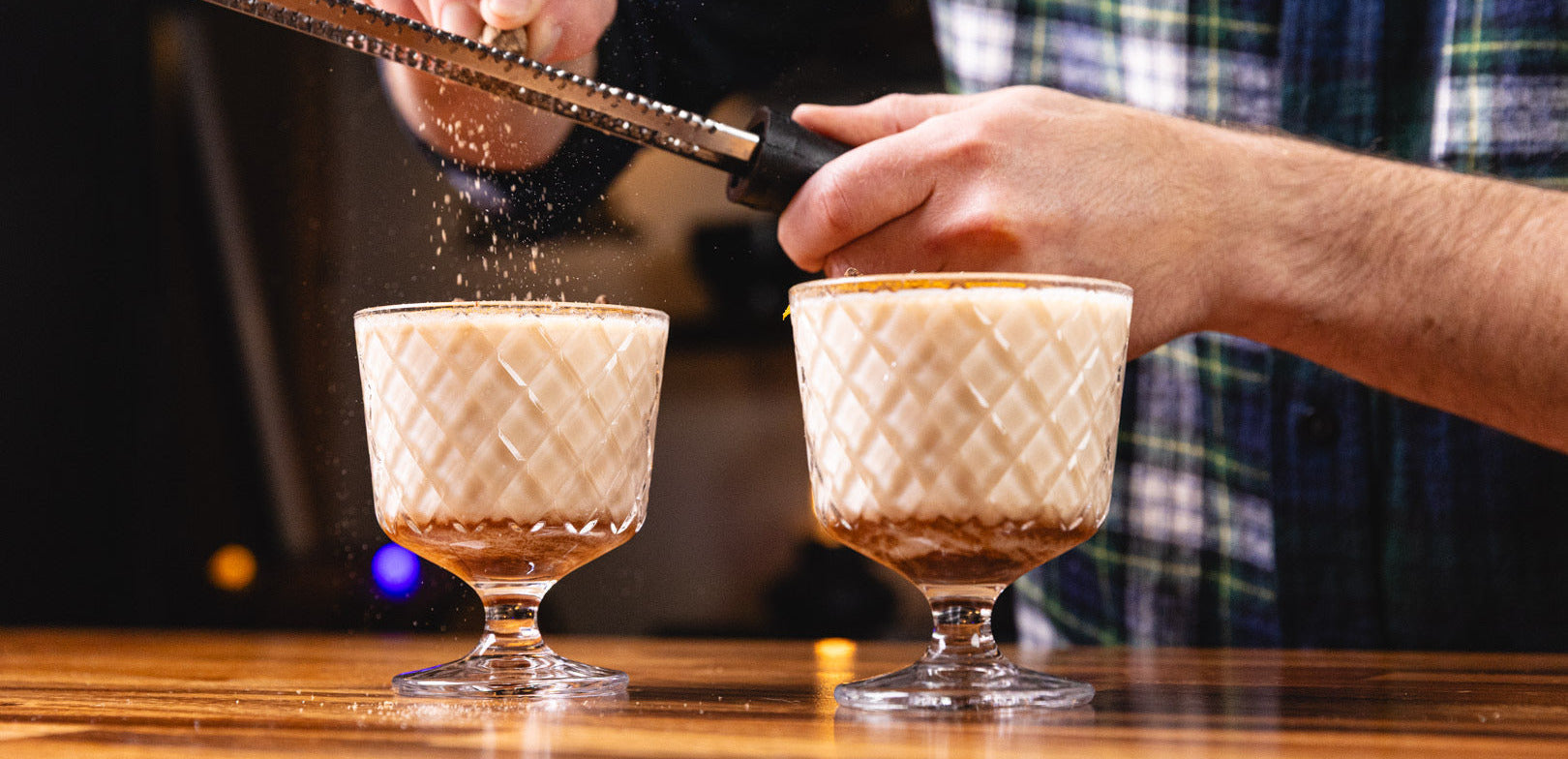 Person garnishing two cocktails with a bar spoon on a wooden bar.